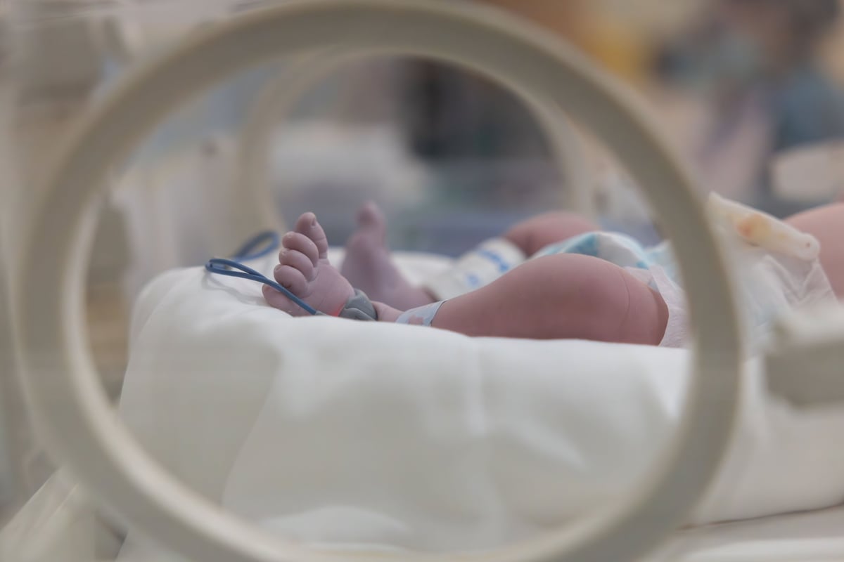 A baby's feet visible in a hospital incubator with blue strap around foot