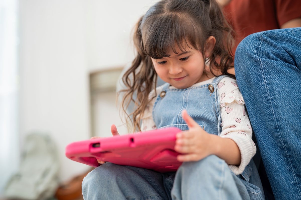 A young child holds a tablet focusing on the screen with an adult beside her