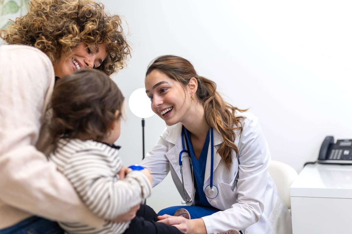 Pediatrician Examining Little Child. Friendly Female Doctor Smiling At Toddler During Medical Checkup. Healthcare And Medicine Concept. Pediatric Care For Baby With Mother In Hospital Office.