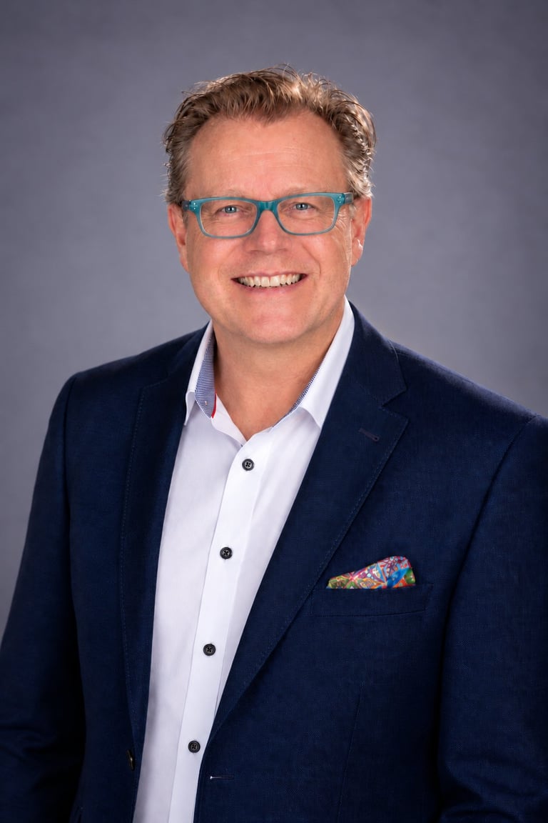 Professional headshot of a smiling man with glasses wearing a navy blazer and white shirt against a gray background