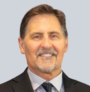 Middle-aged man with brown hair and mustache wearing a dark blazer and checkered shirt with blue tie against light blue background